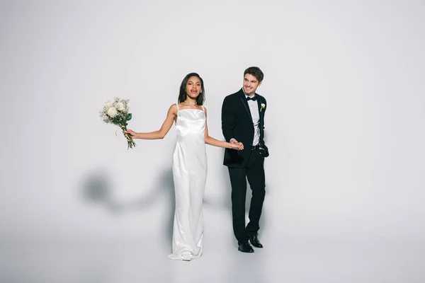 Vue pleine longueur de mariée afro-américaine élégante avec bouquet de mariage tenant la main avec marié heureux sur fond blanc — Photo de stock