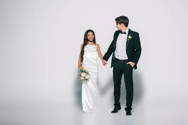Piena vista lunghezza dello sposo bello con mano in tasca che si tiene per mano con bella sposa afro-americana su sfondo bianco — Foto stock