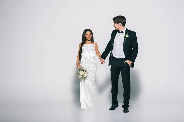 Vue pleine longueur de beau marié avec la main dans la poche tenant la main avec belle mariée afro-américaine sur fond blanc — Photo de stock