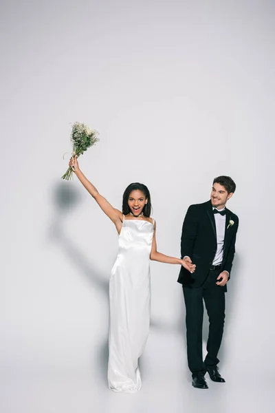 Visão de comprimento total da noiva americana africana feliz segurando buquê de casamento na mão levantada, segurando as mãos com o noivo elegante no fundo branco — Fotografia de Stock