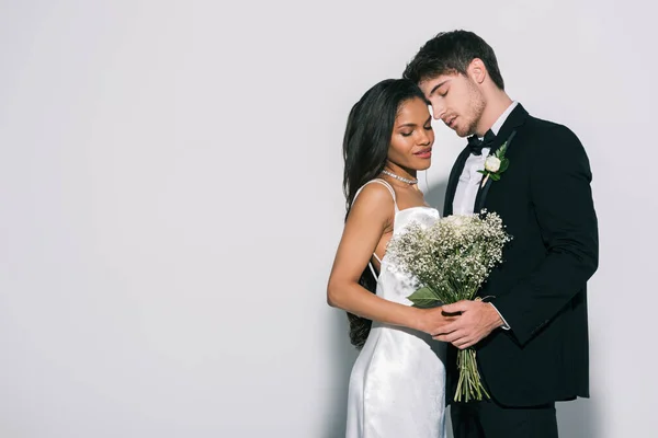 Beau marié et belle mariée afro-américaine avec bouquet de mariage debout face à face avec les yeux fermés sur fond blanc — Photo de stock