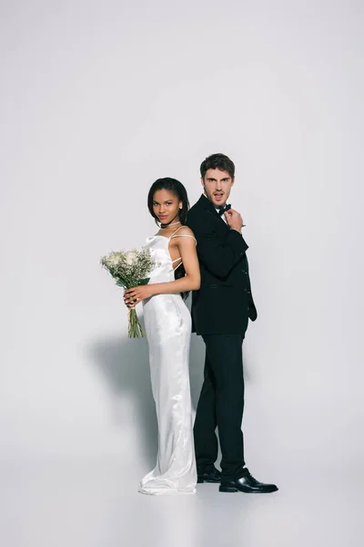 Vista completa de jóvenes, elegantes recién casados interracial de pie espalda con espalda mientras posan sobre fondo blanco - foto de stock