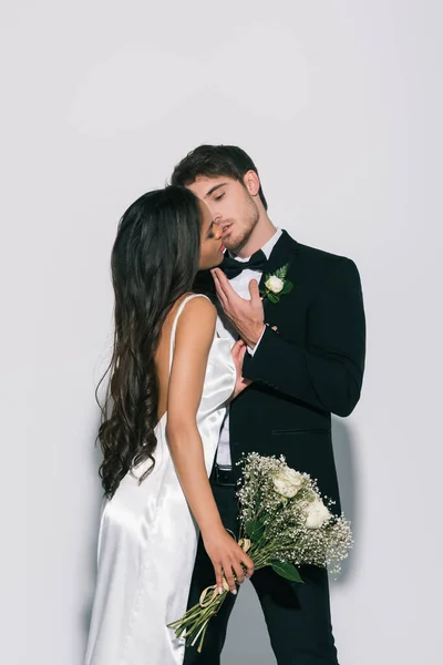 Young bridegroom touching face and kissing beautiful african american bride on white background — Stock Photo