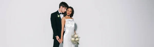 Imagen horizontal de los recién casados interracial elegante de pie con los ojos cerrados sobre fondo blanco - foto de stock
