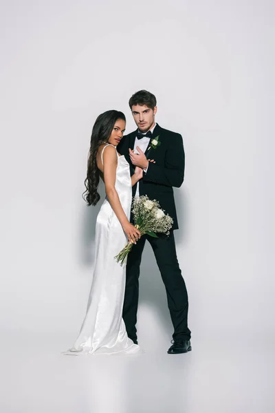 Vue pleine longueur de marié élégant et belle mariée afro-américaine regardant la caméra tout en posant sur fond blanc — Photo de stock