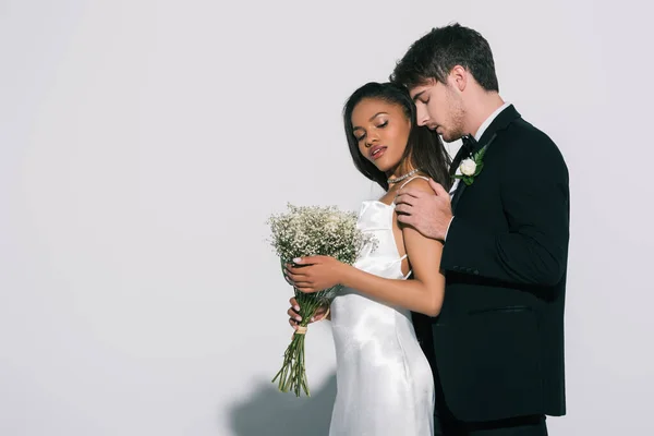 Beau marié touchant les épaules de belle mariée afro-américaine sur fond blanc — Photo de stock