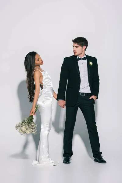 Full length view of elegant interracial newlyweds holding hands and looking at each other on white background — Stock Photo