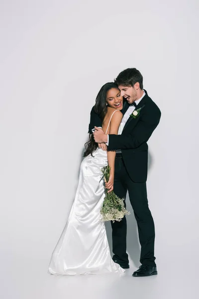 Piena vista lunghezza dello sposo felice abbracciando sorridente sposa africana americana su sfondo bianco — Foto stock