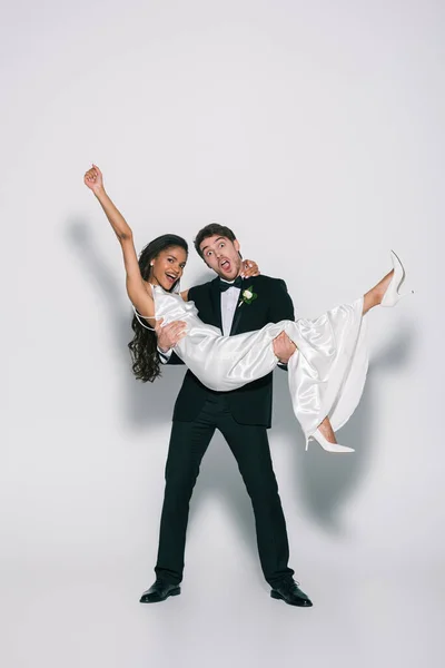 Vue pleine longueur du marié joyeux tenant heureuse mariée afro-américaine sur les mains sur fond blanc — Photo de stock