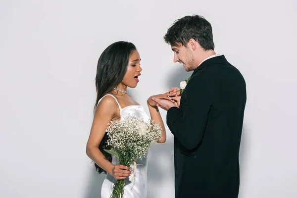 Noivo jovem colocando anel de casamento no dedo da noiva americana africana animado no fundo branco — Fotografia de Stock