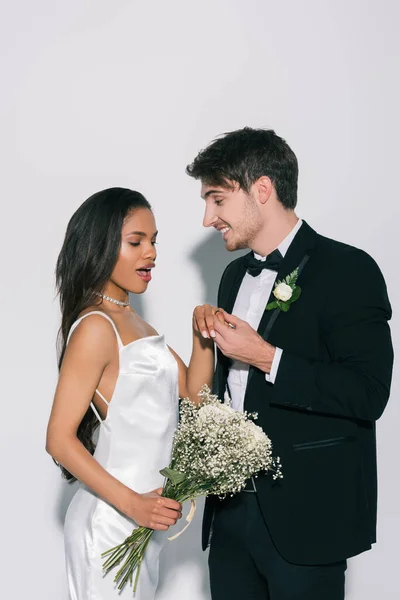 Happy groom holding hand of excited african american bride on white background — Stock Photo