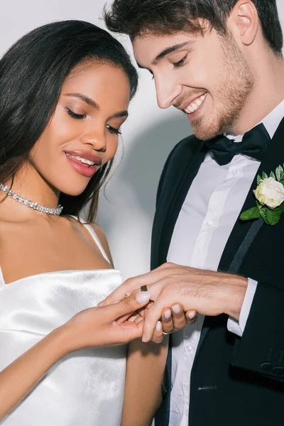 Beautiful african american bride putting wedding ring on finger of happy groom on white background — Stock Photo