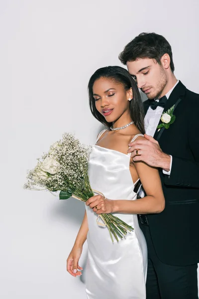 Bello sposo toccando spalla di bella sposa afro-americana tenendo bouquet da sposa su sfondo bianco — Foto stock