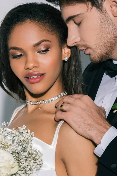 Novio guapo con los ojos cerrados tocando hombro de hermosa novia afroamericana sobre fondo blanco - foto de stock