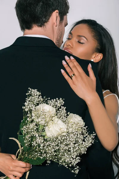 Schöne afrikanisch-amerikanische Braut umarmt Bräutigam, während Hochzeitsstrauß isoliert auf weiß — Stockfoto
