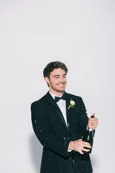 Cheerful bridegroom opening bottle near splashes of champagne on white background — Stock Photo