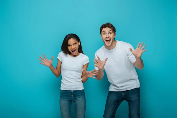 Joyeux couple interracial montrant des gestes effrayants à la caméra sur fond bleu — Photo de stock