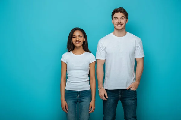 Felice uomo con mano in tasca e allegra ragazza afroamericana sorridente alla macchina fotografica su sfondo blu — Foto stock