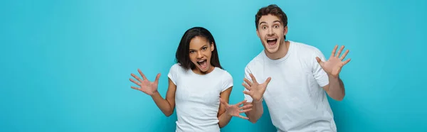 Panoramic shot of cheerful interracial couple showing frightening gestures at camera on blue background — Stock Photo