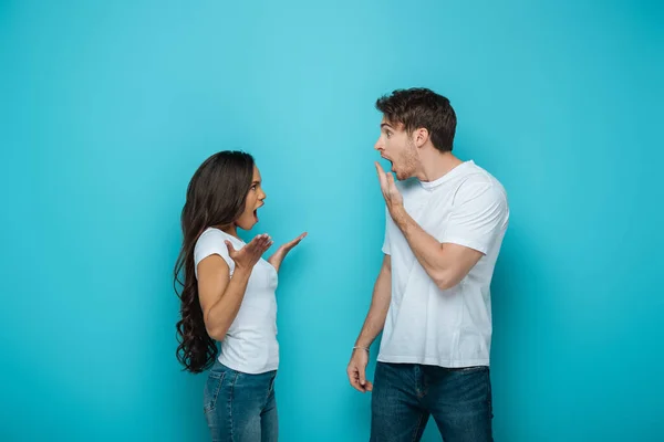 Uomo scioccato che copre la bocca con mano vicino afro-americano ragazza litigando su sfondo blu — Foto stock