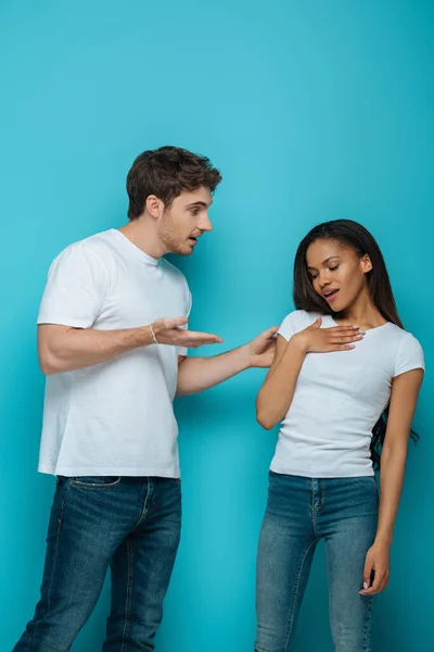 Joven hablando con ofendido afroamericano novia tocando pecho sobre fondo azul - foto de stock