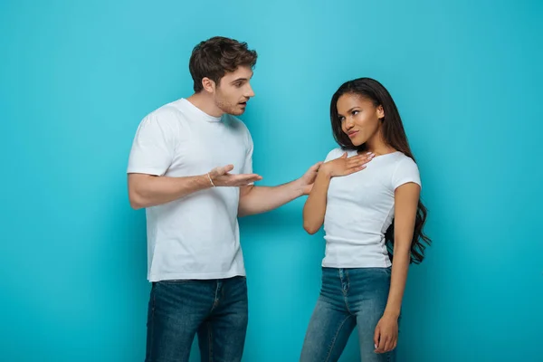 Young man speaking to offended african american girlfriend touching chest on blue background — Stock Photo