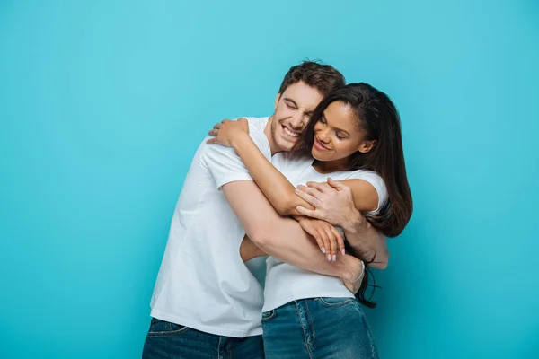 Jovem, feliz casal interracial em t-shirts brancas abraçando com olhos fechados no fundo azul — Fotografia de Stock