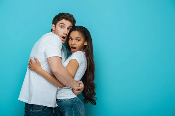 Casal interracial assustado abraçando e olhando para a câmera no fundo azul — Fotografia de Stock