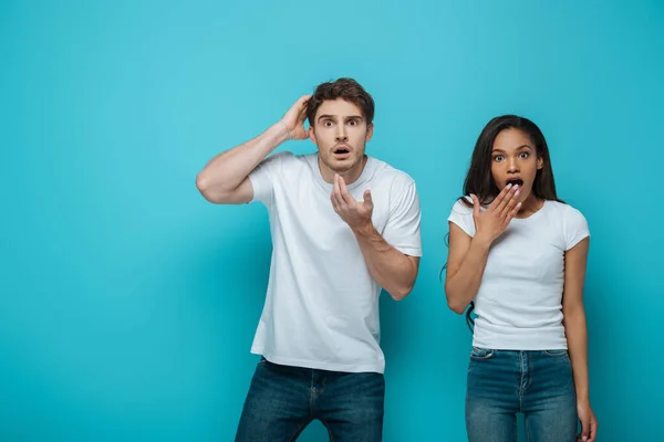 Sorprendido hombre y africano americano chica mirando cámara en azul fondo - foto de stock