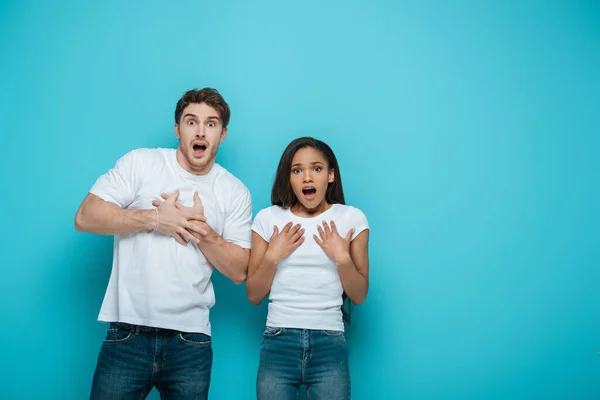 Scared interracial couple touching chests while looking at camera on blue background — Stock Photo