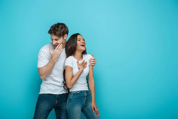 Junger Mann berührt Schulter eines afrikanisch-amerikanischen Mädchens beim gemeinsamen Lachen auf blauem Hintergrund — Stockfoto