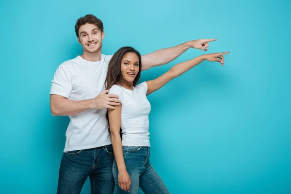 Smiling interracial couple pointing with fingers while looking at camera on blue background — Stock Photo