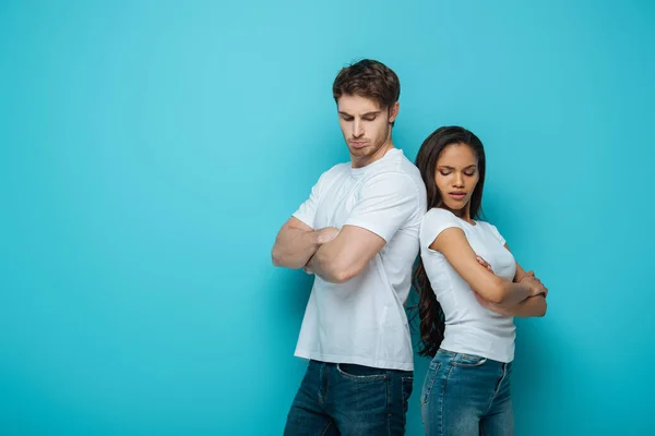 Offended interracial couple standing back to back with crossed arms on blue background — Stock Photo
