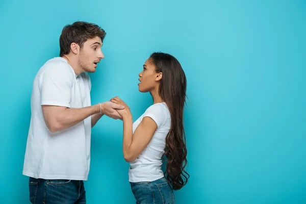 Vista laterale del giovane uomo che si tiene per mano di attraente ragazza afro-americana su sfondo blu — Foto stock