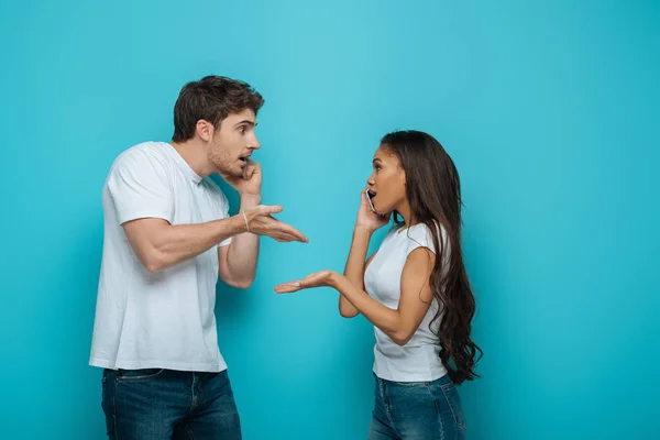 Vista lateral de la joven pareja interracial peleando mientras habla en teléfonos inteligentes sobre fondo azul - foto de stock