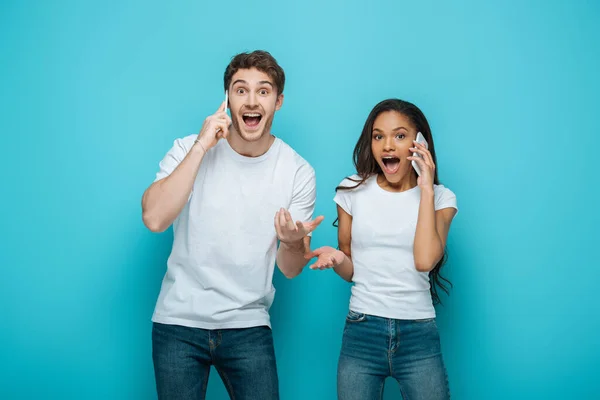 Excited interracial couple talking on smartphones while standing with open arms on blue background — Stock Photo