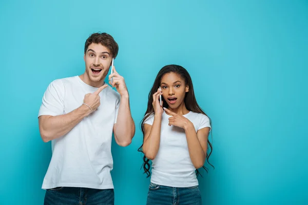 Sorprendido pareja interracial señalando con los dedos mientras habla en los teléfonos inteligentes sobre fondo azul - foto de stock