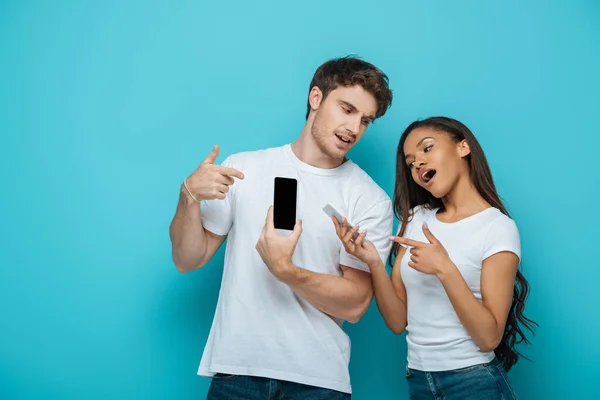 Atractiva chica afroamericana con novio guapo señalando con los dedos a los teléfonos inteligentes sobre fondo azul - foto de stock