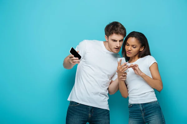 Зосереджена міжрасова пара дивиться на смартфон в руках афроамериканської дівчини на синьому фоні — стокове фото