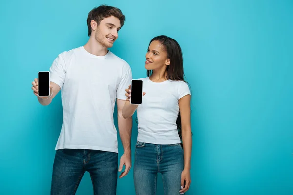 Sonriente pareja interracial mirándose mientras muestra teléfonos inteligentes con pantalla en blanco sobre fondo azul - foto de stock