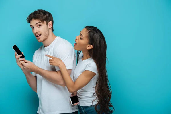 Shocked african american girl pointing with finger at smartphone of boyfriend on blue background — Stock Photo