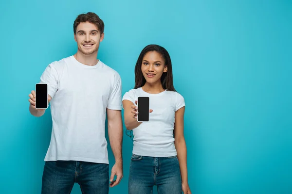 Sonriente pareja interracial mostrando teléfonos inteligentes con pantalla en blanco sobre fondo azul - foto de stock