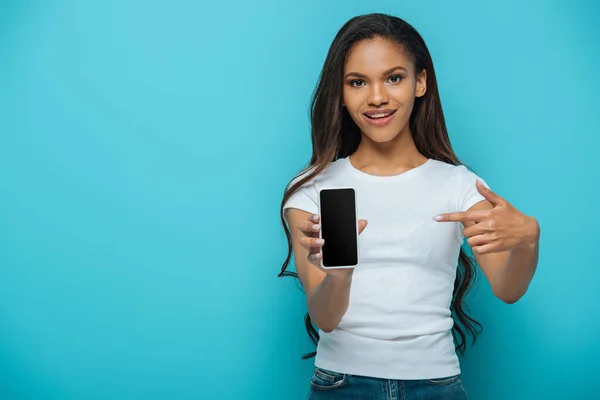 Chica afroamericana sonriente apuntando con el dedo al teléfono inteligente con la pantalla en blanco aislado en azul - foto de stock