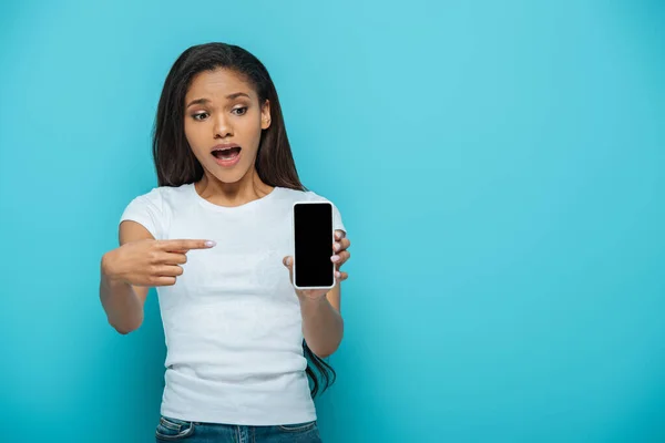 Шокована афроамериканська дівчина вказує пальцем на смартфон з порожнім екраном ізольовано на синьому — стокове фото