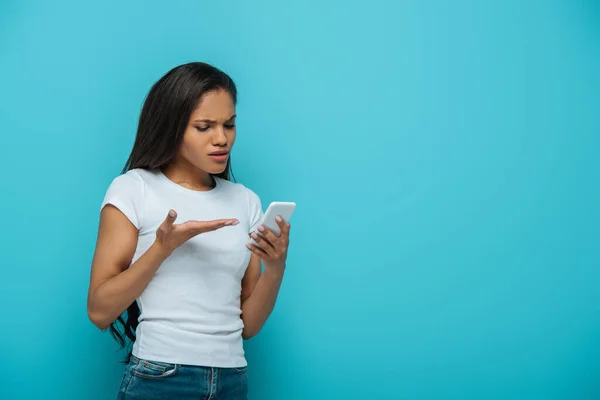 Недовольная африканская американская девушка, стоящая с открытой рукой, глядя на смартфон, изолированный на голубом — стоковое фото