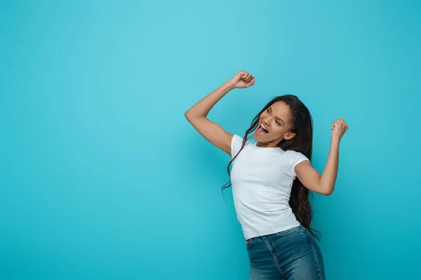 Happy african american girl showing winner gesture while looking away on blue background — Stock Photo