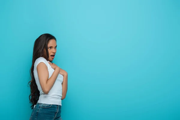 Conmocionada chica afroamericana tocando el pecho mientras mira hacia otro lado aislado en azul - foto de stock