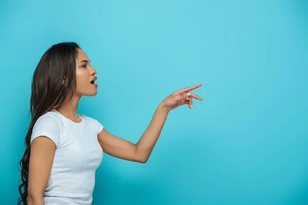 Vue latérale de surprise afro-américaine fille pointant du doigt tout en regardant loin sur fond bleu — Photo de stock