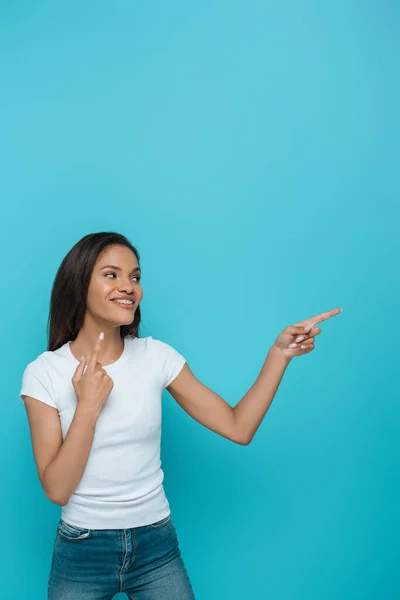 Sonriente afroamericana chica apuntando a los frenos dentales en sus dientes y apuntando lejos aislado en azul - foto de stock