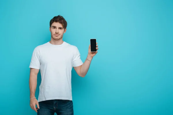 Jeune homme mécontent montrant smartphone avec écran blanc sur fond bleu — Photo de stock